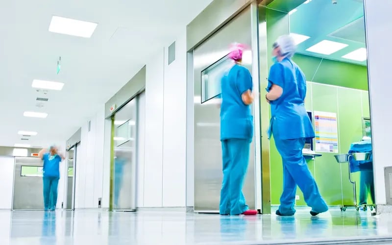 Pflegekräfte in blauer Dienstkleidung gehen durch einen hellen Krankenhausflur.