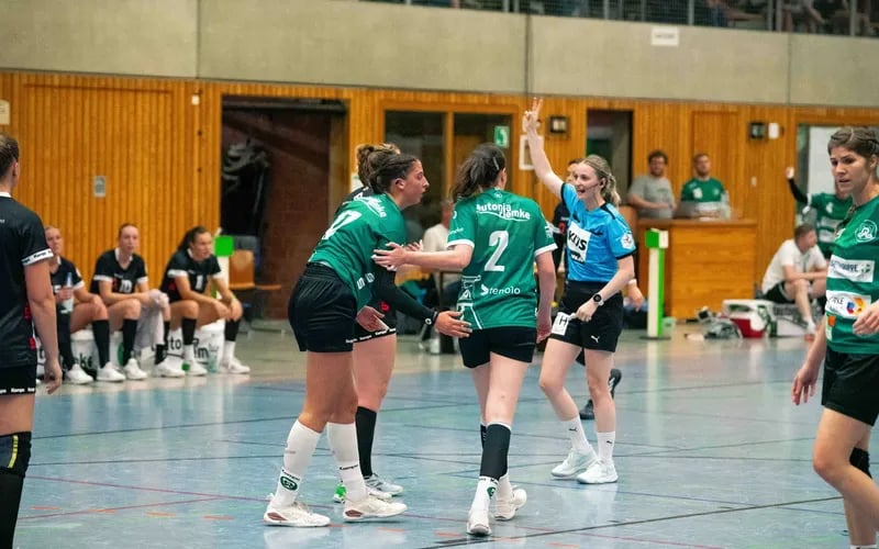 Handballerinnen im grün-weißen Trikot klatschen sich während eines Spiels in der Halle ab.