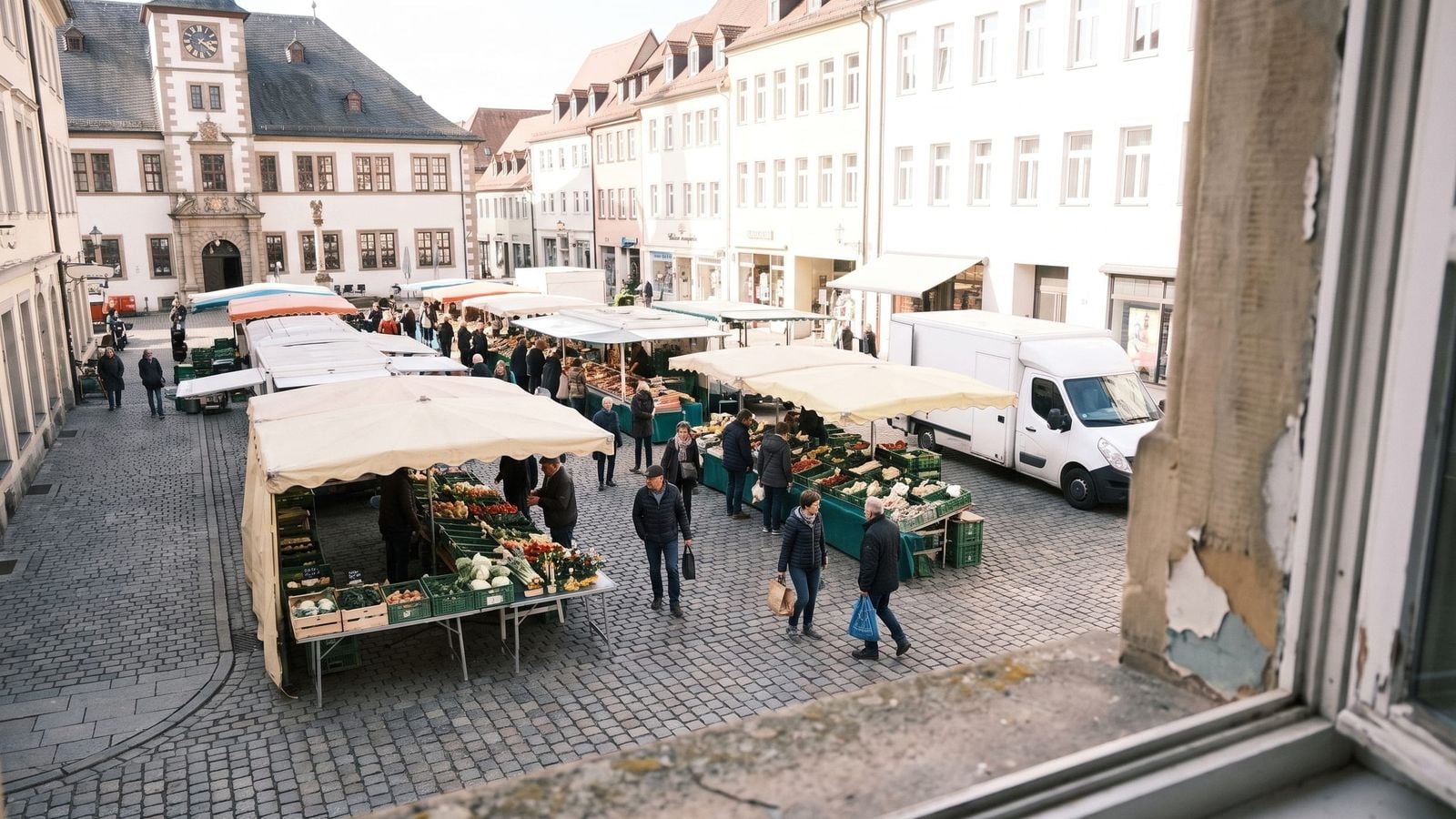 Ein Marktplatz am Vormittag mit der Fassade des Rathauses, einer Reihe Marktstände unter hellen Markisen und Menschen zwischen den Ständen